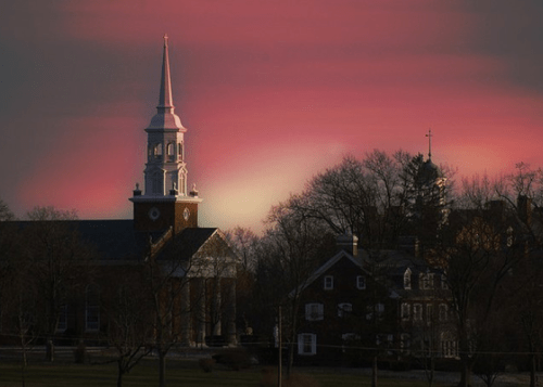 A church steeple silhouetted against a vibrant pink and purple sunset, with trees and buildings in the background.