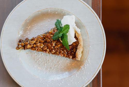 A slice of pecan pie topped with whipped cream and a mint leaf, served on a decorative plate with powdered sugar.