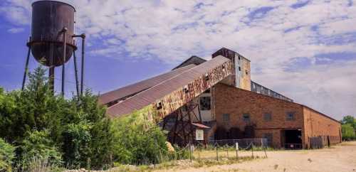 An old industrial building with a sloped roof, water tower, and overgrown vegetation under a cloudy sky.