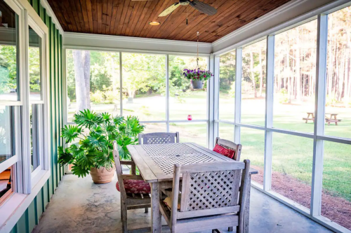 A screened porch with a wooden table and chairs, surrounded by greenery and a view of a grassy yard.
