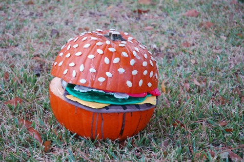 A creatively carved pumpkin designed to look like a hamburger, with sesame seeds and colorful toppings, on grass.