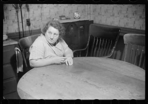 A woman sits at a wooden table in a dimly lit room, looking thoughtfully to the side. Vintage decor in the background.