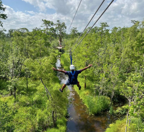 A person zip-lining over a lush green landscape with trees and a winding stream below.