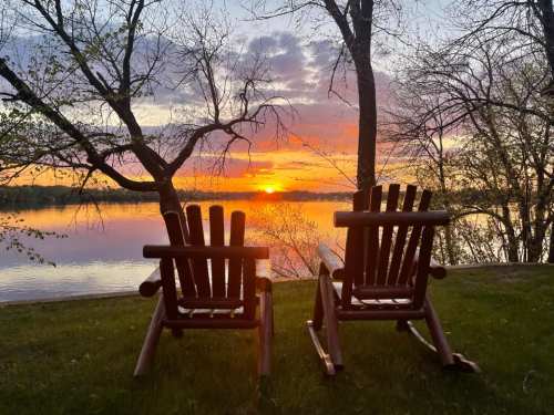 Two wooden chairs facing a vibrant sunset over a calm lake, surrounded by trees and greenery.