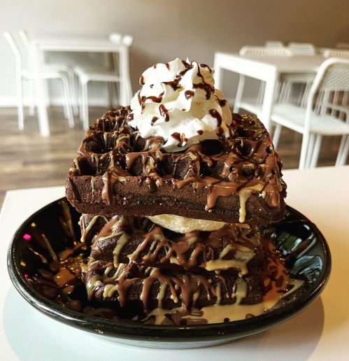 A stack of chocolate waffles topped with whipped cream and drizzled with chocolate and caramel sauces on a black plate.