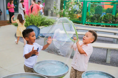 Two young boys joyfully play with a large soap bubble at an outdoor activity area, surrounded by greenery and other children.