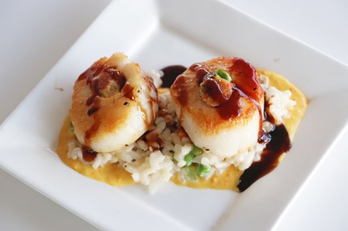 Two seared scallops on a bed of risotto, drizzled with a dark sauce, served on a white square plate.