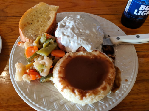 A plate with meatloaf topped with gravy, mashed potatoes with brown gravy, mixed vegetables, and a slice of bread.