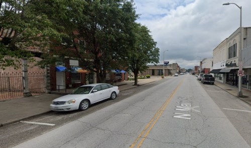 A quiet street scene with trees, parked cars, and buildings lining the road under a cloudy sky.