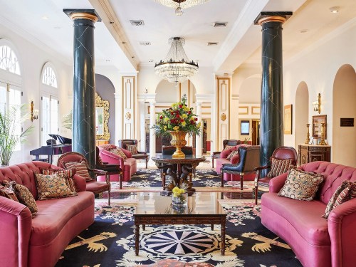 Elegant hotel lobby featuring pink sofas, floral decor, a chandelier, and decorative pillars.