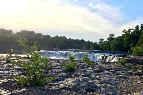 A serene waterfall cascades over rocks, surrounded by lush greenery and a person standing nearby in the sunlight.