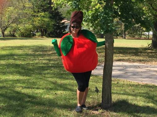 A person in a large red apple costume poses playfully next to a tree in a sunny park.