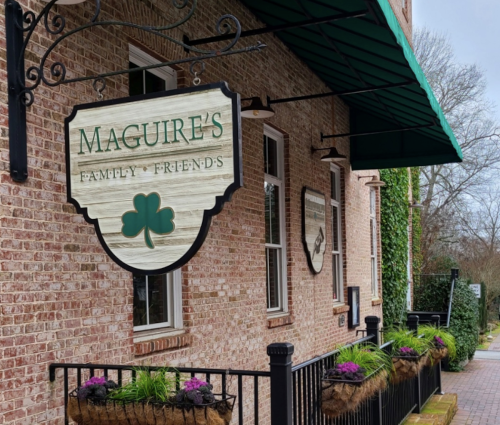 Sign for "Maguire's Family & Friends" with a green shamrock, located on a brick building with flower planters.