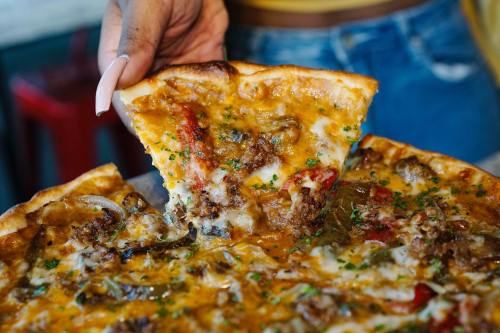 A hand holding a slice of cheesy pizza topped with meat and vegetables, with the whole pizza in view.