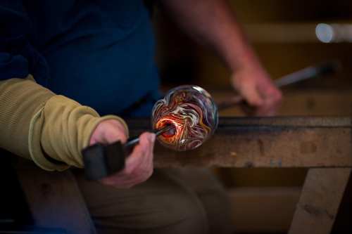 A glassblower shapes a colorful glass piece, holding a tool and working at a wooden bench.