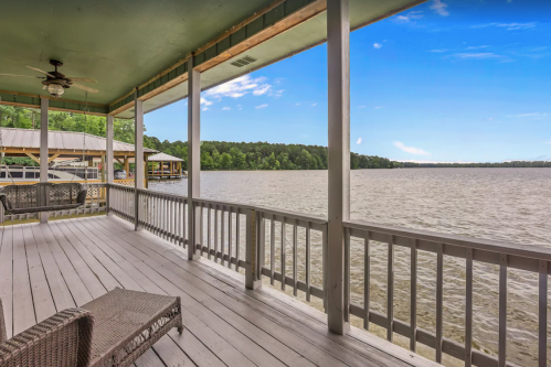 A serene lakeside view from a porch with wooden flooring and a ceiling fan, surrounded by lush greenery.