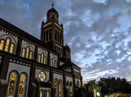 A historic church with stained glass windows, set against a cloudy twilight sky. Palm trees are visible in the foreground.
