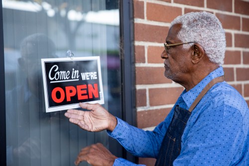An older man in an apron hangs an "Open" sign on a shop window, smiling as he prepares to welcome customers.