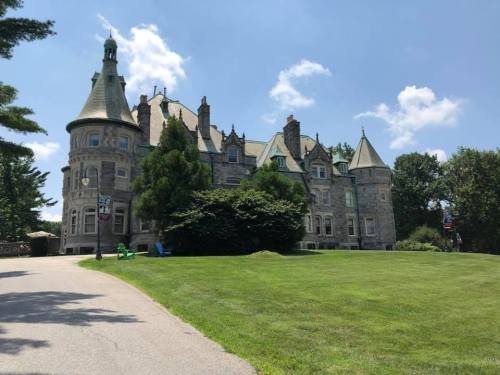 A large, historic stone mansion with turrets, surrounded by green grass and trees under a blue sky.