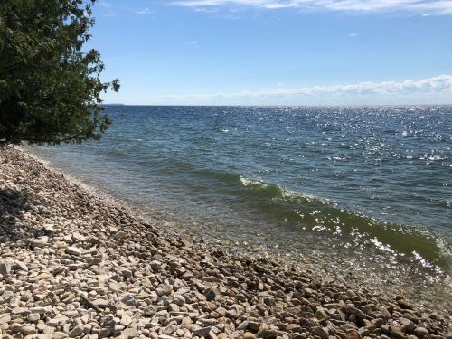 A serene lakeshore with smooth stones, gentle waves, and a clear blue sky in the background.