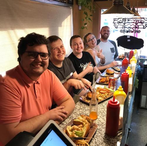 A group of five friends enjoying a meal at a diner, smiling with food and condiments on the table.