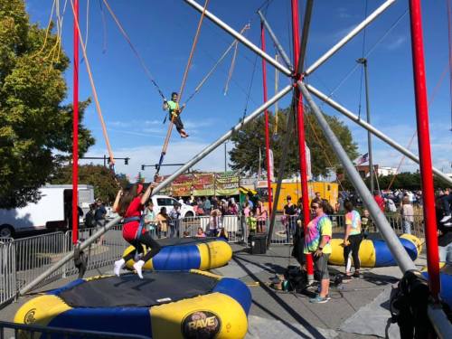 A lively outdoor scene with people enjoying a bungee trampoline setup, surrounded by colorful attractions and spectators.