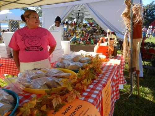 A woman in a pink shirt stands at a table with pies, surrounded by autumn decorations at a market.