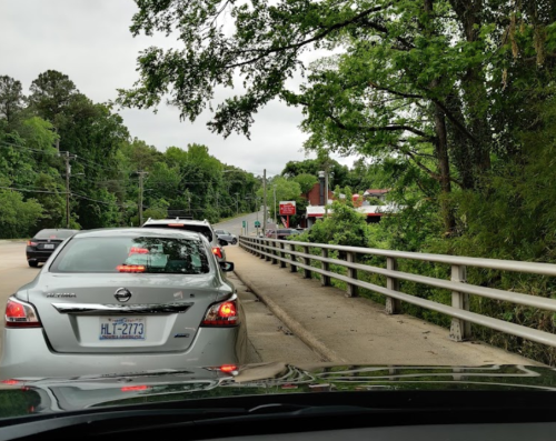 Traffic lined up on a road beside a bridge, with trees and a building visible in the background.