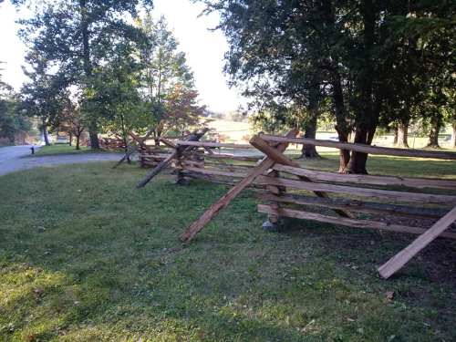 A rustic wooden fence with several sections leaning, surrounded by trees and grass in a peaceful outdoor setting.