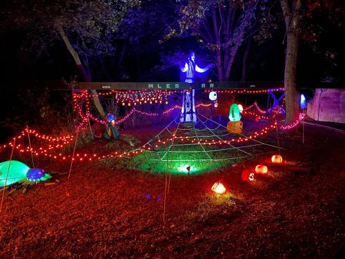A spooky Halloween display with colorful lights, a ghost figure, pumpkins, and spider webs in a dark outdoor setting.