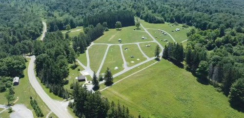 Aerial view of a green campsite with tents and trees, surrounded by a winding road and lush forest.