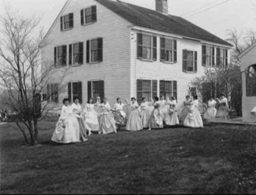 A group of women in vintage dresses poses in front of a large white house with shutters and a garden.