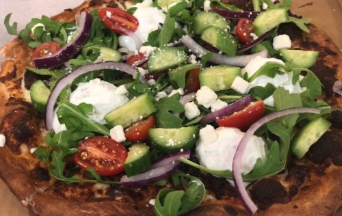 A fresh pizza topped with arugula, cherry tomatoes, cucumbers, red onion, feta cheese, and poached eggs.