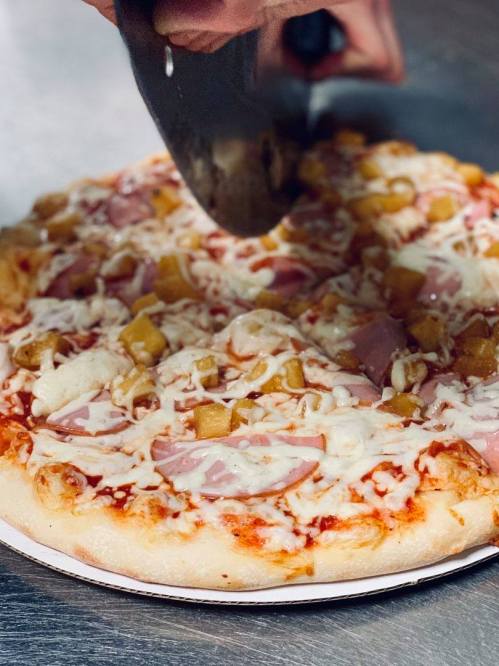 A hand using a pizza cutter to slice a Hawaiian pizza topped with cheese, ham, and pineapple.