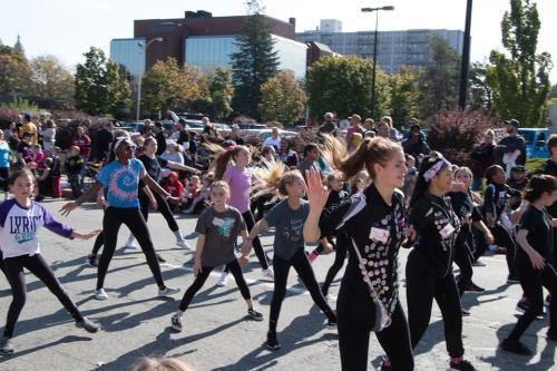 A group of young dancers performing in a lively outdoor setting, with spectators watching in the background.