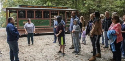 A group of people listens to a guide near a vintage trolley in a wooded area.