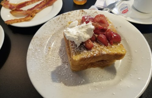 A plate of French toast topped with strawberries and whipped cream, dusted with powdered sugar, alongside crispy bacon.