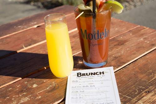 A glass of orange juice and a large cocktail with a lime slice, alongside a brunch menu on a wooden table.