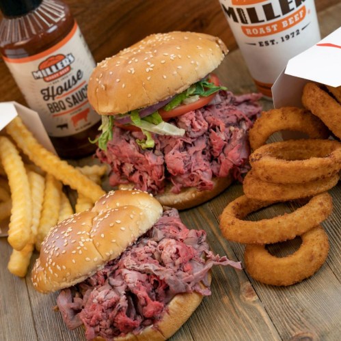 Two large roast beef sandwiches with sesame seed buns, accompanied by fries, onion rings, and BBQ sauce.
