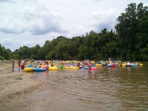 A lively river scene with people floating on colorful inner tubes, surrounded by trees and a sandy shore.