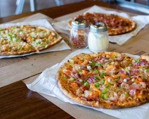 Three pizzas on a table, topped with various ingredients, alongside shakers of red pepper flakes and grated cheese.