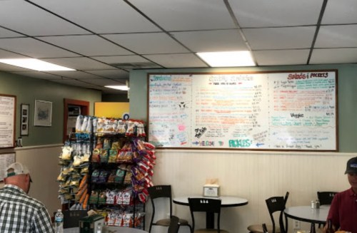 A casual eatery with a menu board on the wall, snack display, and two patrons sitting at tables.