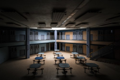 Abandoned cafeteria with empty tables and dim lighting, surrounded by faded walls and closed doors.