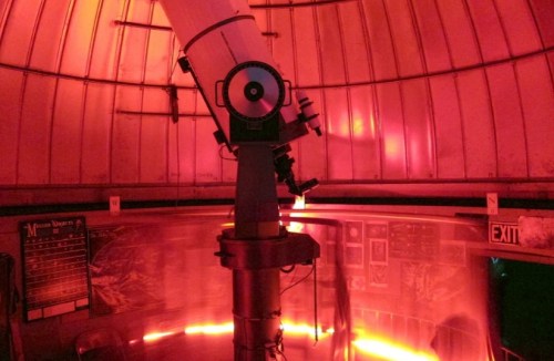 A telescope inside a dimly lit observatory with red lighting and various charts on the walls.