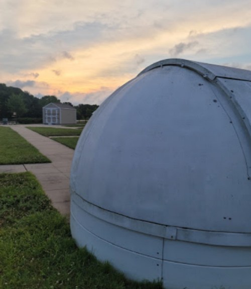 A small, white dome structure sits on a grassy area with a pathway, surrounded by trees and a building in the background.