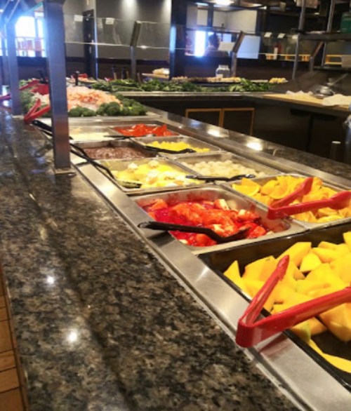 A salad bar with various fresh vegetables and fruits in metal trays on a granite countertop.
