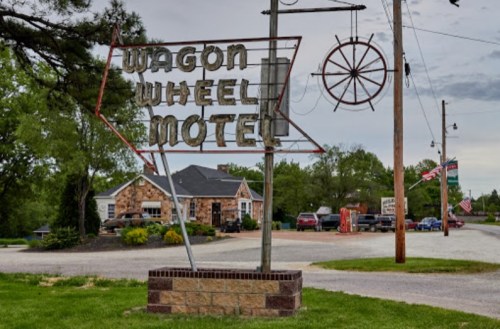 Sign for Wagon Wheel Motel with a rustic design, surrounded by greenery and a few parked cars in the background.