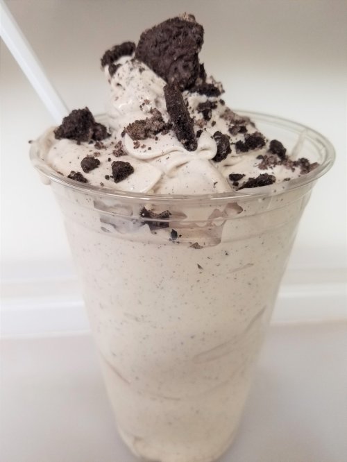 A creamy milkshake topped with whipped cream and crushed chocolate cookies in a clear cup.