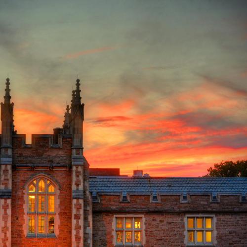 A historic building with gothic architecture against a vibrant sunset sky, featuring warm glowing windows.