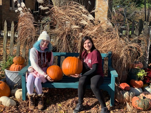 Two young women sit on a teal bench holding pumpkins, surrounded by autumn foliage and decorative pumpkins.
